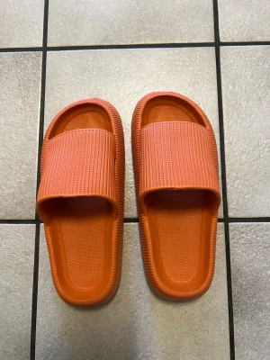 Orange chunky sandaler med bred rem storlek 38-39 - Säljer ett par orangea sandaler med bred ribbad rem och chunky sula. De är tillverkade i mjukt och flexibelt syntetmaterial som är lätt att rengöra. Perfekta för att glida runt hemma eller på stranden. Färgen är riktigt poppig och designen är enkel men trendig.