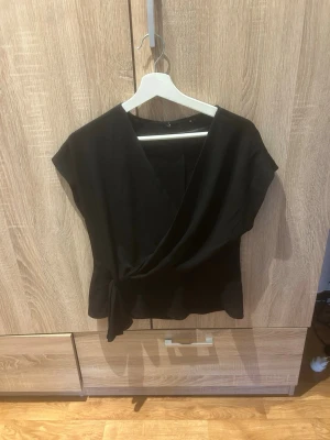 Svart omlottopp med knyt - Stilren svart omlottopp med knyt i sidan och v-ringad hals. Toppen är ärmlös och har en smickrande passform som passar perfekt till både jeans och kjol. Tillverkad i mjukt tyg som känns bekvämt mot huden.