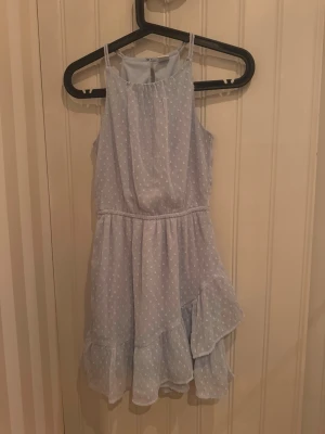 Ljusblå volangklänning - Söt ljusblå kortklänning med vita prickar och volangdetaljer. Klänningen har tunna axelband, är ärmlös och har en luftig passform. Perfekt för varma dagar och somriga tillfällen. Materialet känns lätt och skönt mot huden.