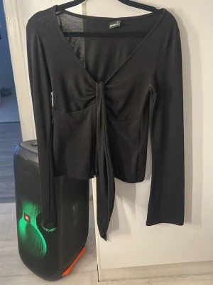 Svart långärmad topp med knyt - Trendig svart långärmad topp med djup v-ringning och snygg knytning framtill. Modellen är croppad och har en smickrande passform som framhäver midjan. Perfekt att styla med höga jeans eller kjol.
