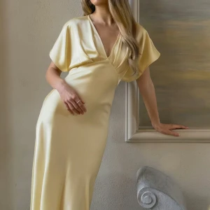 Ljusgul långklänning med v-ringning - Elegant ljusgul långklänning med djup v-ringning och korta, vida ärmar. Klänningen har en mjuk, följsam passform och ett stilrent, minimalistiskt uttryck. Perfekt för dig som gillar en feminin och tidlös look.