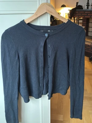 Mörkgrå cropped kofta med knappar - Snygg mörkgrå cropped kofta med rund halsringning och knäppning framtill. Långa ärmar och ribbade muddar vid halsen ger en clean look. Perfekt att slänga över en topp för en chill outfit.