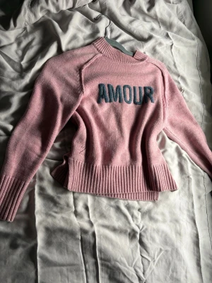 Rosa stickad tröja Zadig & Voltaire - Söt rosa stickad tröja från Zadig & Voltaire med blå texten 'AMOUR' framtill. Tröjan har ribbade muddar vid hals, ärmslut och nederkant samt en avslappnad passform. Perfekt för dig som gillar trendiga och bekväma plagg.