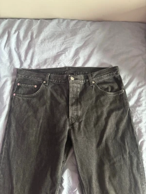 Svarta raka jeans från Weekday W-32 L-34 - Säljer ett par svarta jeans från Weekday med klassisk femficksdesign och raka ben. Jeansen har silverfärgade nitar och knapp. Tillverkade i denim med en snygg tvättad finish. Storlek W-32 L-34