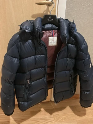 Svart dunjacka - Snygg svart dunjacka med huva och tvåvägsdragkedja. Jackan har fickor med dragkedja framtill och en extra ficka med Moncler-logga på ärmen. Klassisk puffer-modell som håller dig varm och stilren under kalla dagar.