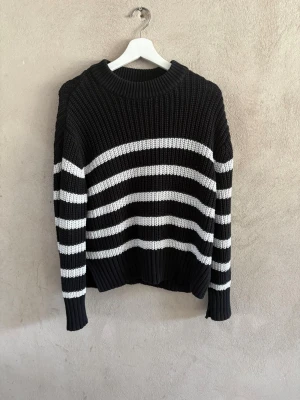Svartvit randig stickad tröja - Säljer en svart stickad tröja med vita horisontella ränder. Tröjan har en rund halsringning, ribbade muddar och lång ärm. Perfekt för dig som gillar en klassisk och avslappnad stil.