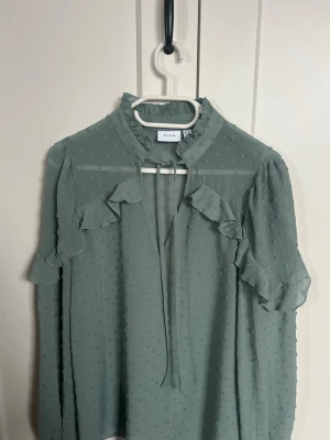 Grön volangblus från VILA - Ljusgrön blus från VILA med långärmad design och volangdetaljer längs axlarna. Blusen är tillverkad i ett tunt, transparent tyg med diskret prickstruktur och har en liten ståkrage med knytband framtill. Perfekt för en trendig och avslappnad look.