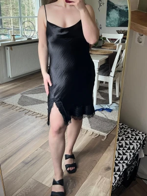 Svart satinklänning med spets - Svart kortklänning i satin med tunna axelband och spetsdetaljer längst ner samt vid slit. Passar även M skulle jag säga. 