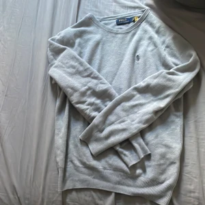 Ljusgrå tröja från Polo Ralph Lauren - Ljusgrå långärmad tröja från Polo Ralph Lauren med rund halsringning och diskret broderad logga på bröstet. Tröjan är i ett tunt, mjukt material och har ribbade muddar vid ärmslut och nederkant. Perfekt för en clean och stilren look.