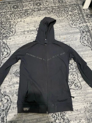 Svart zip hoodie från Nike - Säljer en svart hoodie från Nike med dragkedja framtill och huva. Hoodien har diskreta mönsterdetaljer, två fickor med dragkedja och en liten patch nertill med texten 'Authentic Nike F.C. 1984'. Perfekt för en sportig och avslappnad stil.