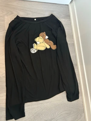 Svart långärmad tröja med nalletryck - Svart långärmad tröja med ett gulligt tryck av två nallar på bröstet, en ljusgul och en brun. Tröjan har rund halsringning och är gjord i ett mjukt, stretchigt material som känns skönt mot huden. Perfekt för dig som gillar söta detaljer och vill ha en unik look.