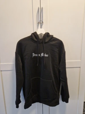 Svart hoodie Dream Maker print - Svart hoodie med huva och magficka. På bröstet står det 'Dream Maker' i vit text och på ryggen finns ett stort tryck med text, grafik och konstmotiv. Hoodien har dragsnören vid huvan och ribbade muddar. Materialet känns mjukt och bekvämt, perfekt för en chill look.