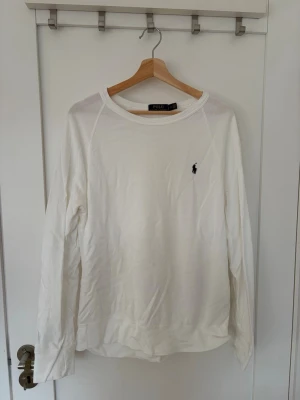 Vit långärmad tröja från Polo Ralph Lauren - Klassisk vit långärmad tröja från Polo Ralph Lauren med rund halsringning och den ikoniska lilla broderade loggan på bröstet. Tillverkad i mjukt bomullstyg som känns skönt mot huden. Perfekt till jeans eller chinos för en clean och stilren look. Tröjan är stor i storlek så skulle säga att den egentligen är L