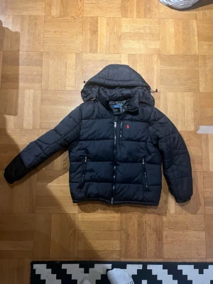 Svart pufferjacka från Polo Ralph Lauren - Snygg svart pufferjacka från Polo Ralph Lauren med röd logga på bröstet. Jackan har huva, två dragkedjeförsedda fickor och quiltad design. Perfekt för kalla dagar och riktigt skön att bära. Klassisk och tidlös stil som funkar till det mesta.