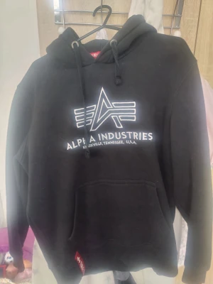 Svart hoodie Alpha Industries - Säljer en svart hoodie från Alpha Industries med stor vit logga och text på bröstet. Hoodien har huva med dragsko, magficka och ribbade muddar. Perfekt för dig som gillar streetwear och vill ha en klassisk look.