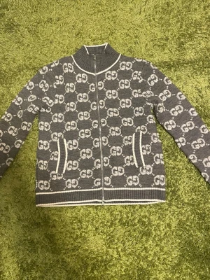 Grå ziptröja från Gucci med GG-mönster - Snygg grå ziptröja från Gucci med ikoniskt GG-mönster över hela tröjan. Tillverkad i 100% ull och har vita detaljer vid dragkedja, krage, fickor och nederkant. Perfekt för dig som vill ha en exklusiv och trendig look.