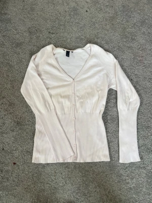 Ljusrosa ribbad kofta med knappar - Säljer en ljusrosa kofta med ribbade detaljer och knäppning framtill. Koftan har långa ärmar, v-ringad hals och smal passform. Perfekt att slänga över en topp för en chill look.