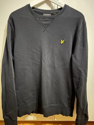 Svart sweatshirt från Lyle & Scott - Klassisk svart sweatshirt från Lyle & Scott med rund halsringning och gul logga broderad på bröstet. Tröjan har ribbade muddar vid ärmslut och nederkant. Perfekt för en avslappnad och stilren look.