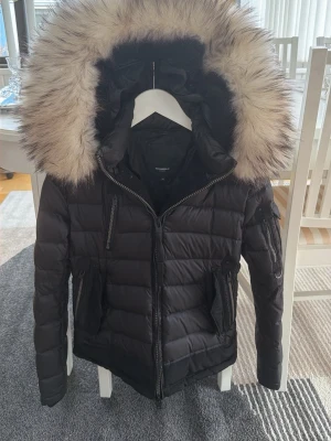 Svart dunjacka Rockandblue med pälshuva - Snygg svart dunjacka från Rockandblue med stor fluffig pälskant på huvan. Jackan har flera dragkedjeförsedda fickor, quiltad design och en varm, vadderad insida. Perfekt för kalla vinterdagar och riktigt trendig look. Kostar ifrån början ca 5000kr 
