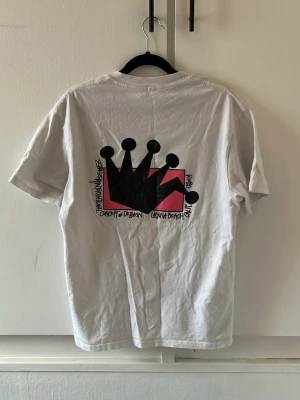 Vit Stüssy t-shirt - Vit t-shirt från Stüssy med ikoniskt svart kronmotiv och logga på bröstet samt stort tryck på ryggen. Klassisk passform och rund halsringning. Tillverkad i mjuk bomull, perfekt för en avslappnad streetwear-look.