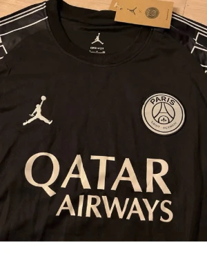 Svart PSG x Jordan matchtröja - Säljer en svart Paris Saint-Germain matchtröja i samarbete med Jordan. Tröjan har vita detaljer, PSG-logga och Jordan-logga på bröstet samt Qatar Airways tryckt på framsidan. Unika mönster på ärmarna och rund hals. Perfekt för fotbollsfans!