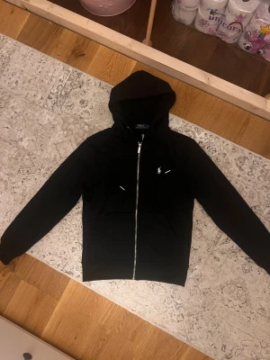 Svart hoodie från Polo Ralph Lauren - Säljer en svart hoodie från Polo Ralph Lauren med dragkedja framtill och klassisk vit logga på bröstet. Hoodien har huva med snören och två fickor framtill. Perfekt för en avslappnad och stilren look.