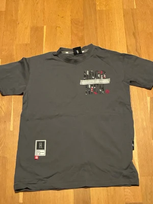 HOODRICH t-shirt - Snygg grå t-shirt från HOODRICH med stort tryck på både fram- och baksidan där det står 'PRODUCT OF THE STREETS'. T-shirten har korta ärmar och camomönster i trycket. Perfekt för dig som gillar streetwear och vill sticka ut.