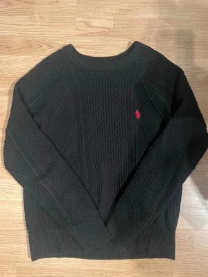 Svart stickad tröja från Ralph Lauren - Klassisk svart stickad tröja från Ralph Lauren med kabelstickat mönster och röd broderad logga på bröstet. Tröjan har rund halsringning och långa ärmar, perfekt för en stilren och tidlös look.
