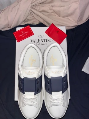 Valentino Garavani Open sneakers vit/blå - Säljer ett par Valentino Garavani Open sneakers i vitt skinn med bred mörkblå rem över snörningen. Snygga detaljer med guldlogga på plösen och innersulan samt mönstrad vit yttersula. Perfekt för dig som gillar lyxiga och stilrena sneakers. (Säljs pga att dom inte passar)