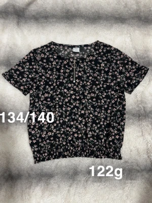  blommig blus med dragkedja - En blus från Lindex med  blommor, korta ärmar och dekorativ dragkedja framtill. Blusen har resår nedtill för en bekväm passform och är perfekt för dig som gillar ett romantiskt och avslappnat uttryck.🌸 Fint använt skick. Storlek 134/140 