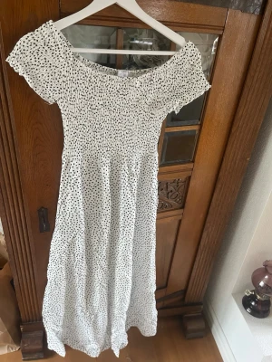 Vit o svart prickig  klänning - Söt vit långklänning med svarta prickar. Klänningen har smock upptill och korta ärmar, vilket ger en avslappnad och trendig look. Perfekt för varma dagar och somriga tillfällen. 