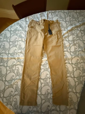 Beige chinos från Tommy Hilfiger - Snygga beige chinos från Tommy Hilfiger med klassisk rak passform. Tillverkade i mjukt bomullstyg och har bälteshällor, knappgylf och sidofickor. Perfekta för en clean och stilren look.