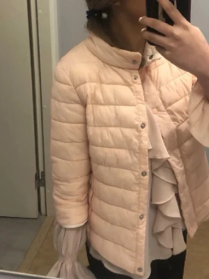 Trekvartärmad jacka  - Säljer en ljusrosa pufferjacka med ståkrage och tryckknappar framtill. Jackan har quiltad design och är lätt vadderad, perfekt för kyliga dagar. Passar dig som gillar en stilren och enkel look. Materialet är syntet och jackan har en kortare modell.