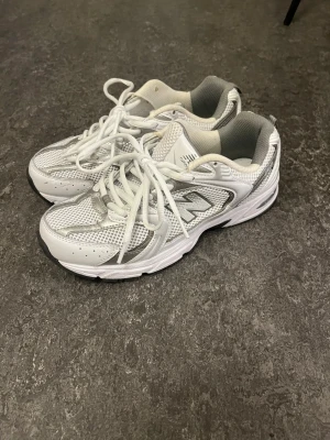 New Balance 530 vita sneakers - Snygga New Balance 530 sneakers i vitt med silverdetaljer och meshpaneler för extra ventilation. Klassisk sportig look med bekväm sula och snörning. Perfekta för dig som gillar retrostil och streetwear.