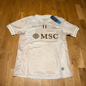 Napoli away kit 25/26 EA7 - Snygg vit matchtröja från Napoli med gulddetaljer och MSC-logga på bröstet. Tröjan har subtila mönster i tyget, italiensk flagga och klubbmärke. Tillverkad i lätt och ventilerande material, perfekt för fotboll eller träning.