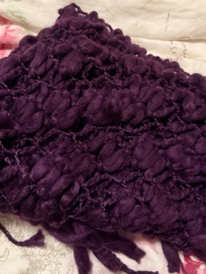 Lila chunky halsduk med tofsar - Unik lila halsduk i chunky stickad design med stora garnbollar och dekorativa tofsar längs kanterna. Halsduken har ett öppet, luftigt mönster och känns riktigt mysig och trendig. Perfekt för dig som vill sticka ut med en färgstark accessoar.