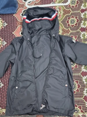 Svart vindjacka med huva Moncler (äkta) (byta) - Snygg svart vindjacka från Moncler med huva och dragsko. Jackan har dragkedja framtill, två fickor med tryckknappar och en cool detalj med röd, vit och blå rand på huvan. Materialet är glansigt och vattenavvisande, perfekt för blåsiga dagar.
