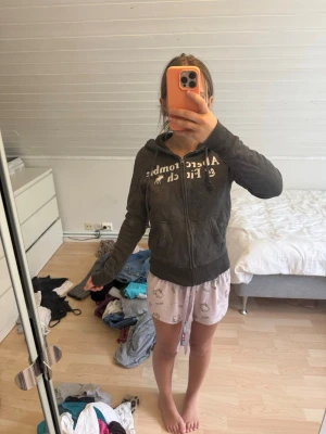 Mörkgrå sipup hoodie från Abercrombie - Mörkgrå sipup hoodie från Abercrombie & Fitch med dragkedja framtill och vit logga på bröstet. Hoodien har huva med dragsko och två fickor framtill. Perfekt för en avslappnad och chill stil. Dragkedjan är lite trög:)