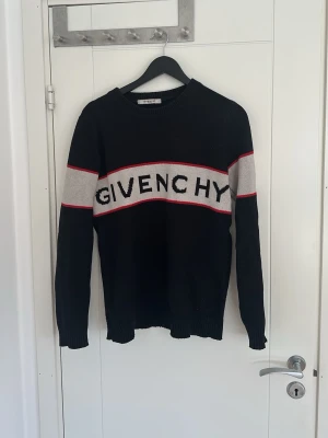 Svart Givenchy tröja med logga sällsynt  - Snygg svart stickad tröja från Givenchy med vit och röd rand över bröstet och armarna samt stor Givenchy-logga framtill. Tröjan har rund halsringning och långa ärmar. Perfekt för dig som gillar streetwear och exklusiva märken.