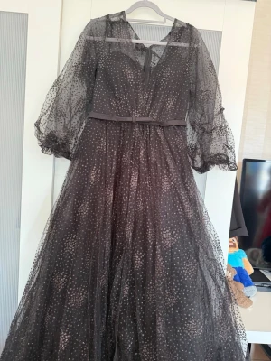 Svart långklänning med glitterprickar - Magisk svart långklänning med transparenta puffärmar och glittriga prickar över hela tyget. Klänningen har v-ringning, knytband vid halsen och ett smalt bälte i midjan. Perfekt för dig som vill sticka ut med en elegant och unik look.