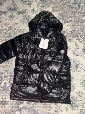 Svart dunjacka med huva från Moncler - Snygg svart dunjacka från Moncler med glansig finish och huva. Jackan har dragkedja framtill, två sidofickor med dragkedja och Moncler-logga på ärmen. Perfekt för kalla vinterdagar och riktigt trendig look.