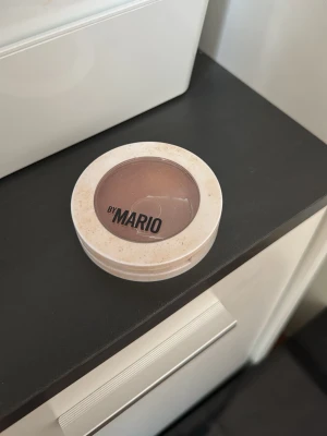 By Mario bronzer i varm brun nyans - Bronzer från By Mario i en rund, beige plastförpackning med transparent lock. Produkten har en varm brun färg som ger en naturlig solkysst look. Perfekt för att framhäva ansiktskonturer och ge extra glow.