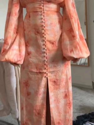 Lång adoore klänning med puffärm - ADOORE Lång klänning i ljus orange och rosa toner med blommigt mönster. Klänningen har puffiga långärmar, knappar framtill och en slits. Snygg passform med markerad midja och lätt glansigt material.