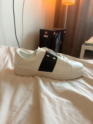 Valentinos vita med svart detalj  - Snygga vita sneakers från Valentino med en bred svart läderdetalj över sidan och silverfärgade nitar. Klassisk låg modell med rund tå och vita skosnören. Perfekta för dig som gillar stilrena och exklusiva sneakers. Bara använda vid provning där jag märke att de var för små.
