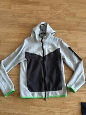 Nike Tech Fleece hoodie grå/svart - Nike Tech Fleece hoodie i ljusgrått med svart panel framtill och vita loggan på bröstet. Hel dragkedja, huva och gröna detaljer vid ärmslut och nederkant. Materialet är mjukt och sportigt, perfekt för en avslappnad streetwear-look.