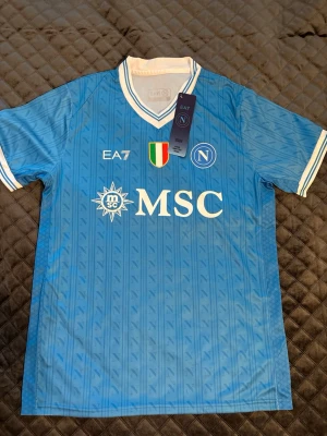 Napoli matchtröja EA7 blå - Snygg Napoli matchtröja från EA7 i ljusblått med vita detaljer. Tröjan har V-ringad krage med dubbla ränder, klubbmärke och sponsortryck på bröstet samt italienska mästerskapsmärket. Tillverkad i lätt och ventilerande material, perfekt för fotboll eller träning.