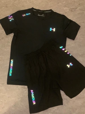 Svart träningsset från Under Armour - Snyggt svart träningsset från Under Armour med t-shirt och shorts. Setet har färgglada, reflekterande loggor och textdetaljer på både tröja och byxa. Tillverkat i ett lätt och snabbtorkande material som passar perfekt till träning och sport.