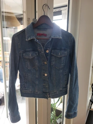 Klassisk blå jeansjacka - Snygg blå jeansjacka med klassisk passform och silverfärgade knappar framtill. Jackan har två bröstfickor med lock och knapp samt markerade sömmar för en tidlös look. Perfekt att slänga över en hoodie eller t-shirt.