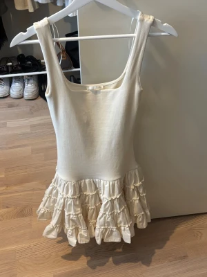 Beige volangklänning med ärmlös design - Söt vit kortklänning med volanger nertill och ärmlös design. Klänningen har en rundad urringning och är tillverkad i ett mjukt bomullstyg som känns bekvämt mot huden. Perfekt för varma dagar och ger en romantisk känsla med sina lager av volanger.