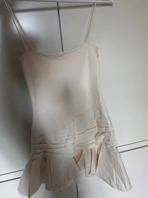 Ljus kort klänning från Zara - Säljer en ljus, kort klänning från Zara med tunna axelband och asymmetrisk volangdetalj nedtill. Klänningen har en enkel, stilren design och är perfekt för varma dagar. 